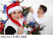 Девушка в шапке Санта-Клауса на фоне молодого человека, наряжающего елку. Стоковое фото, фотограф Phovoir Images / Фотобанк Лори