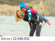 Молодая девушка весело катает на спине маленькую девочку на берегу водоема в прохладную погоду. Стоковое фото, фотограф Иванов Алексей / Фотобанк Лори