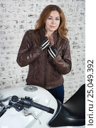 Curly hair attractive woman taking off her leather brown jacket, standing in garage with motorcycle, brick wall background. Стоковое фото, фотограф Кекяляйнен Андрей / Фотобанк Лори