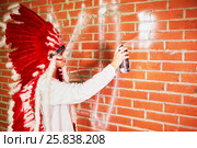 Young woman dressed in costume made of red and white feathers paints on the brick wall with spray. Стоковое фото, фотограф Losevsky Pavel / Фотобанк Лори