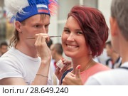 Волонтер в шапке с российской национальной символикой кисточкой рисует на щеке улыбающейся девушки национальный флаг. Москва, Россия, Никольская улица. Чемпионат мира по футболу FIFA 2018. Редакционное фото, фотограф Наталья Николаева / Фотобанк Лори