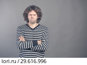 Young man with dark brown hair wears black and white striped casual tshirt looks angry, lips pursed frowning brows, isolated over grey background. Keeps arms crossed and looks at camera. Disagreement. Стоковое фото, фотограф Marina Sharova / Фотобанк Лори