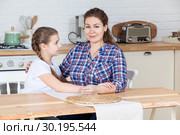 Mother and her ten years old daughter sitting together at table in kitchen, woman looking at camera, girl embracing her mom. Стоковое фото, фотограф Кекяляйнен Андрей / Фотобанк Лори