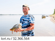 Только что пойманного крупного сазана рыбак держит в руках на фоне реки (2019 год). Редакционное фото, фотограф Игорь Низов / Фотобанк Лори