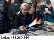 Женщина-стеклодув обрабатывает стеклянную заготовку огнем в мастерской на фабрике ёлочных игрушек в поселке Крестцы Новгородской области (2019 год). Редакционное фото, фотограф Румянцева Наталия / Фотобанк Лори