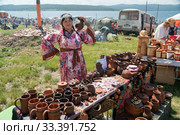 Продавщица с глиняным кувшином в руках стоит за прилавком с посудой на фестивале музыки и ремёсел "Каратаг". Красноярский край (2019 год). Редакционное фото, фотограф Светлана Попова / Фотобанк Лори