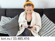 Stay at home. Social distancing. Woman at home relaxing on sofa couch drinking tea from white cup, listening to relaxing music, stay connected to friens and family via social networks on mobile. Стоковое фото, фотограф Matej Kastelic / Фотобанк Лори