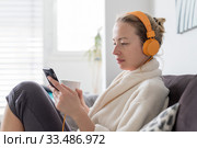Stay at home. Social distancing. Woman at home relaxing on sofa couch drinking tea from white cup, listening to relaxing music, stay connected to friens and family via social networks on mobile. Стоковое фото, фотограф Matej Kastelic / Фотобанк Лори