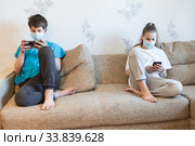 Preteen boy and girl playing online with smartphones while staying at home, kids wearing medical masks, keeping distance. Стоковое фото, фотограф Кекяляйнен Андрей / Фотобанк Лори