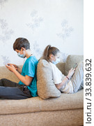 Pre-teen children gaming online using cellphones while sitting at home, kids wearing facial masks, sitting back to back on couch. Стоковое фото, фотограф Кекяляйнен Андрей / Фотобанк Лори
