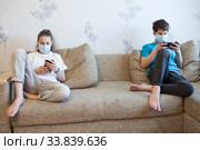 Children keep distance while using smartphones, kids wearing facial mask at home during coronavirus pandemic. Стоковое фото, фотограф Кекяляйнен Андрей / Фотобанк Лори