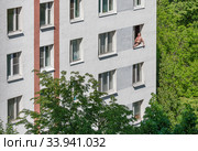 Во время самоизоляции мужчина в плавках загорает, сидя в распахнутом окне многоэтажного жилого дома высоко над землей (2020 год). Редакционное фото, фотограф Наталья Николаева / Фотобанк Лори
