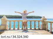 Rear view of happy woman on vacation, wearing straw summer hat ,standing on luxury elegant old stone balcony of coastal villa, relaxing, arms rised to the sun, looking at blue Adriatic sea. Стоковое фото, фотограф Matej Kastelic / Фотобанк Лори