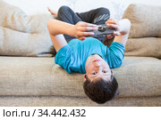Attractive youngster guy wearing blue t-shirt lying on his back on couch and holding smartphone in hands, browsing social media. Стоковое фото, фотограф Кекяляйнен Андрей / Фотобанк Лори