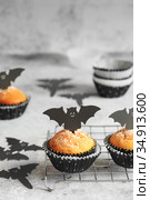 Halloween pumpkin muffins in black capsules decorated with cardboard bats. Festive Halloween cupcakes. Close-up view of delicious spooky halloween muffins on the grey table. Halloween recipes. Стоковое фото, фотограф Nataliia Zhekova / Фотобанк Лори