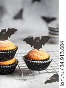 Halloween pumpkin muffins in black capsules decorated with cardboard bats. Festive Halloween cupcakes. Close-up view of delicious spooky halloween muffins on the grey table. Halloween recipes. Стоковое фото, фотограф Nataliia Zhekova / Фотобанк Лори