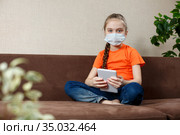 Little girl in medical mask sitting on the sofa in the Lotus position and using tablet pc. Isolation at home. Looking into the camera. Стоковое фото, фотограф Иван Карпов / Фотобанк Лори