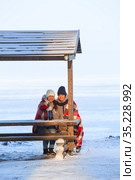 Two frozen girlfriends wrapping with red scarf sitting on wooden bench and drinking hot beverages from thermo mugs for warming up, winter sea beach. Стоковое фото, фотограф Кекяляйнен Андрей / Фотобанк Лори