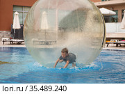 Little boy have fun inside big plastic balloon on the water of swimming pool on the summer resort. Kid inside big inflatable transparent ball running and having fun. Стоковое фото, фотограф Nataliia Zhekova / Фотобанк Лори