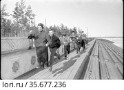 Несколько молодых людей бегут по деревянной площадке над зрительскими трибунами на стадионе «Буревестник», тренировка спортсменов под предводительством мастера из Москвы, Якутия, апрель 1959. Редакционное фото, фотограф Варфоломей Тетерин / Фотобанк Лори