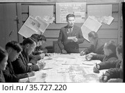 Портрет агитатора, стоящего во главе стола, несколько мужчин сидят на переднем плане и записывают под его диктовку, стенд "Читайте у нас" на заднем плане, Томпо, Якутия, 1965. Редакционное фото, фотограф Варфоломей Тетерин / Фотобанк Лори