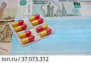 Упаковка с желто-красными капсулами антибиотика на фоне одноразовой медицинской маски и российских банкнот. Стоковое фото, фотограф Цибаев Алексей / Фотобанк Лори
