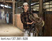 Asian female riding instructor walking in stable with saddle in hands. Стоковое фото, фотограф Яков Филимонов / Фотобанк Лори
