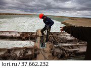 Small Aral Sea Kok-aral dam. Worker in orange hardhat with safety belt and chain use steel rope to lifts water shutter weighting agent. Second worker's hand (right). (2012 год). Редакционное фото, фотограф Alexey Rezvykh / Фотобанк Лори