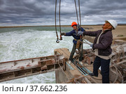 Kok-aral, Kazakhstan - May 03, 2012: Small Aral Sea Kok-aral dam. Hydraulic engineer and worker prepair to lift water shutter weighting agent. Crane's rope with hooks. Stormy sky and turbulent water. Редакционное фото, фотограф Alexey Rezvykh / Фотобанк Лори