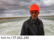 Kok-aral, Kazakhstan - May 03, 2012: Aral Sea Kok-aral dam. Portrait of senior Asian hydraulic engineer on turbulent water river and storm clouds background. Редакционное фото, фотограф Alexey Rezvykh / Фотобанк Лори
