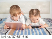 Дети и гаджеты. Мальчик с планшетом и девочка с телефоном лежат на кровати. Стоковое фото, фотограф Владимир Ходатаев / Фотобанк Лори