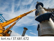 Aktau, Kazakhstan - May 19, 2012 Construction of asphaltic bitumen plant refinery column. Metal distillation tower and yellow hydraulic hoist. Blue sky with clouds. Редакционное фото, фотограф Alexey Rezvykh / Фотобанк Лори