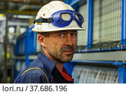 Pavlodar, Kazakhstan - May 29 2012: Caustic chemical plant. Chemical engineer in white hardhat with blue protective glasses on electrolytic process machine background. Looks straight in camera. Редакционное фото, фотограф Alexey Rezvykh / Фотобанк Лори