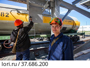 Caustic chemical plant. Two workers in orange hardhat, green protective glasses, blue workwear painting metal stairway on grey and yellow railway tank wagon bkgd (2012 год). Редакционное фото, фотограф Alexey Rezvykh / Фотобанк Лори