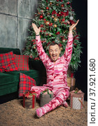 Cheerful surprised funny man in pink sleepwear with hands up sitting near decorated fir tree and Christmas present. Стоковое фото, фотограф Darkbird77 / Фотобанк Лори