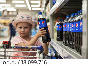 St Petersburg, Russia, july 2022: Adorable little girl sits in shopping cart takes buying pepsi cola on store shelf. Portrait little kid going shopping in store or supermarket. Copy text space. Редакционное фото, фотограф Aleksandr Golubev / Фотобанк Лори