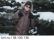 Portrait smiling brutal young man in open air among snow covered Christmas trees in winter season in forest. Tourist with glasses and backpack got lost in an unfamiliar area. Teenage crazy walks. Стоковое фото, фотограф Aleksandr Golubev / Фотобанк Лори