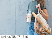 Современная стильная женщина со стеклянной бутылкой молока и авоськой купила хлеб. Девушка на фоне серой стены. Концепция здорового питания и экологии. Отказ от пластиковых пакетов. Стоковое фото, фотограф Photojuli / Фотобанк Лори