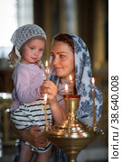 Russian beautiful woman in a scarf and with red hair holding a little girl and lights a candle in front of an icon in the Russian Orthodox Church. Стоковое фото, фотограф Арзюков Вадим Николаевич / Фотобанк Лори
