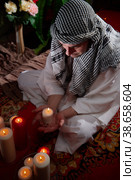 Arab sheikh in a white robe and a headscarf in a boudoir in a room with a red carpet and candles with smoke. Oriental eastern fabulous style photo shoot with a man. Male model posing in fary tale. Стоковое фото, фотограф Кривошеина Елена Леонидовна / Фотобанк Лори
