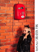 Little girl model in leather jacket calling old red telephone at brick wall background, looking away. Child pretty lady talking payphone outdoor. Communication lifestyle concept. Copy text space. Стоковое фото, фотограф Aleksandr Golubev / Фотобанк Лори