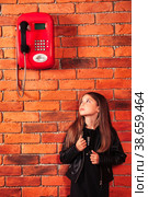 Funny little girl looking on old red payphone at brick wall background. Small brunette lady model in leather jacket looking up on telephone. Communication concept. Copy space for advertising text. Стоковое фото, фотограф Aleksandr Golubev / Фотобанк Лори