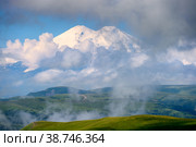 Great nature mountain range. Amazing perspective of caucasian snow mountain or volcano Elbrus with cloudy green field, blue sky background. Elbrus landscape view - highest peak of Russia and Europe. Стоковое фото, фотограф Евгений / Фотобанк Лори