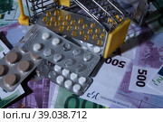 medicines and pills are lying in a small shopping cart against the background of money euro dollars 100 500. pills are poured out of the cart for money the problem of the cost of medicines expensive m. Стоковое фото, фотограф Meshkov Sergey / Фотобанк Лори