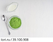 Top view of organic detox matcha ground powder of green tea leaves served in glass bowl with plant milk and spoon on white wooden background with copy space used as ingredient of healthy latte drink. Стоковое фото, фотограф Елена Кабенкина / Фотобанк Лори