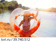 Girl in a hat stands on the river bank with a transparent inflatable circle in the shape of a heart with orange feathers inside. Beach holidays, swimming, tanning, sunscreens. Стоковое фото, фотограф Ольга Симонова / Фотобанк Лори