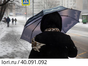 Женщина закрывает лицо зонтом от порывов ветра и мокрого снега на улице во время снегопада в спальном районе города Москвы, Россия (2023 год). Редакционное фото, фотограф Николай Винокуров / Фотобанк Лори