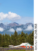 Beautiful rural landscape of Tunka foothill valley with rustic wooden house against backdrop of spruce forest and Eastern Sayan Mountains with low clouds on sunny summer day. Siberia. Buryatia. Arshan. Стоковое фото, фотограф Виктория Катьянова / Фотобанк Лори