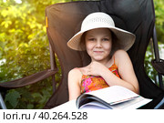 Девушка в соломенной шляпке сидит в походном кресле, держа книгу на коленях, и улыбается. Чтение в летней школе. Стоковое фото, фотограф Юлия Мелешина / Фотобанк Лори