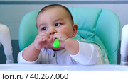 A hungry baby is gnawing on a plastic spoon at the table on a high chair. Teething, whims, itchy gums, introduction of complementary foods. Стоковое видео, видеограф Ольга Симонова / Фотобанк Лори